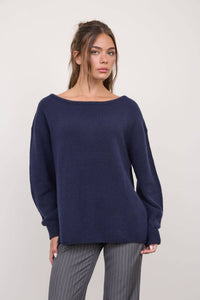A woman is wearing The Workshop Studio’s Boat Neck Oversized Knit Pullover Sweater in Red Wood, paired with gray pinstripe pants. She stands against a plain light background, looking at the camera with a neutral expression.