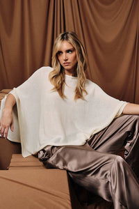 A woman with long blonde hair is wearing the Sunday Edition by LE LIS - LOOSE FIT OPEN KNIT SWEATER CAPE TOP in brown, size large, as she sits on a brown draped sofa with tan fabric hanging in the background.
