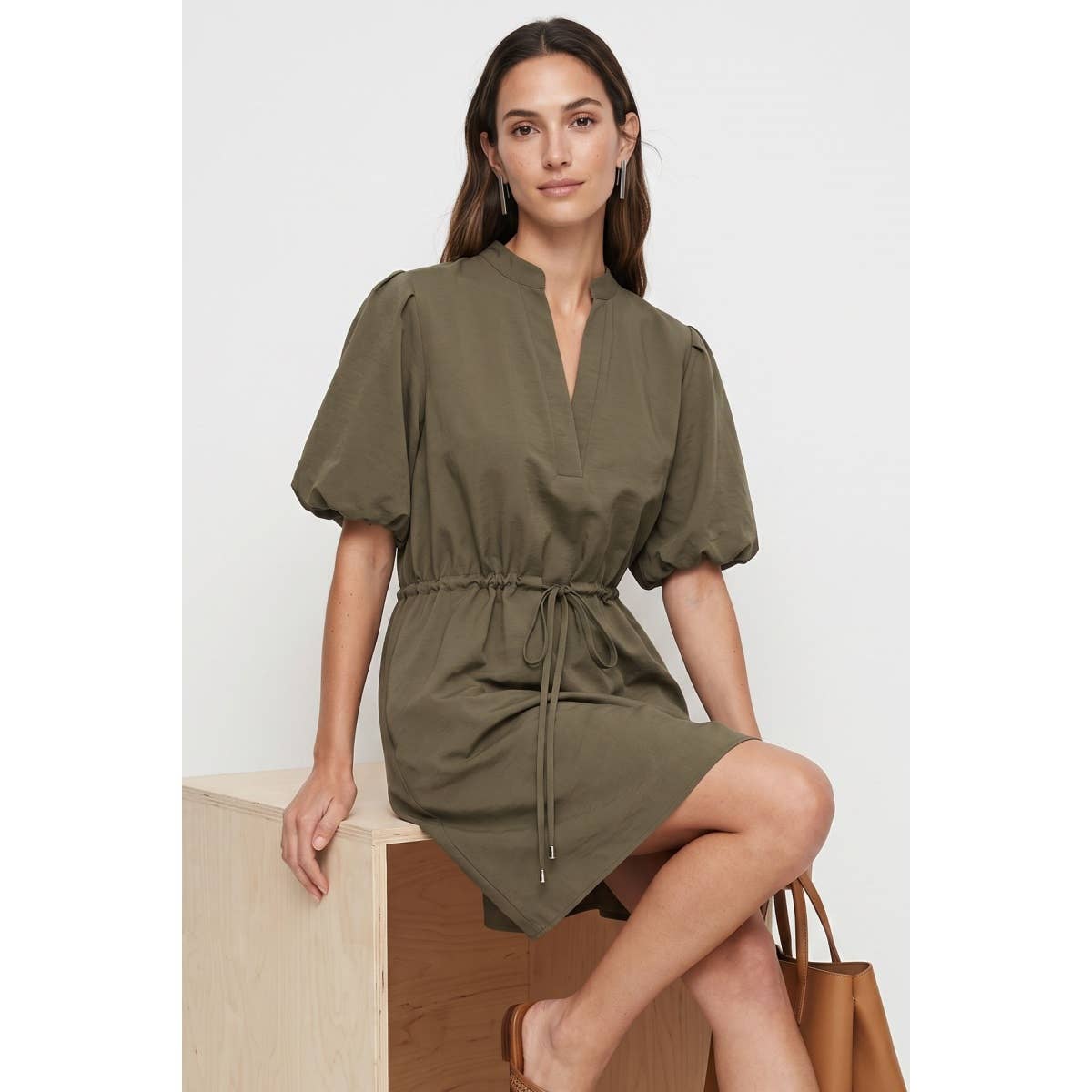 A woman wearing a Naked Zebra - Puff Sleeve Cinched Waist Mini Dress: Navy / L in olive green with a V-neck and a drawstring waist, sitting on a wooden cube.