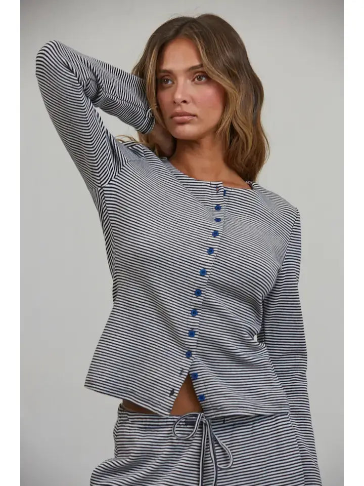 A woman in the Creamon - CTP3592 BROWN Lounge Striped Button Down Top & Pants Set, featuring brown stripes, an asymmetrical button-down top, and matching wide leg pants, stands with one hand behind her head, gazing off to the side.