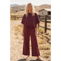 A woman stands on a dirt path by a fence, dressed in the Heather Brushed Stretch Boxy Top & Wide Leg Pant Set—a cozy loungewear ensemble in maroon featuring a loose boxy top and matching wide-leg pants—paired with tan shoes. Hills, grass, and a barn appear in the background beneath a clear sky.