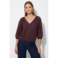 A woman wears a burgundy Naked Zebra - Elastic Puff Sleeve V-neck Blouse: Burgundy / L with gathered puff sleeves and an elasticized waist, paired with dark blue jeans.