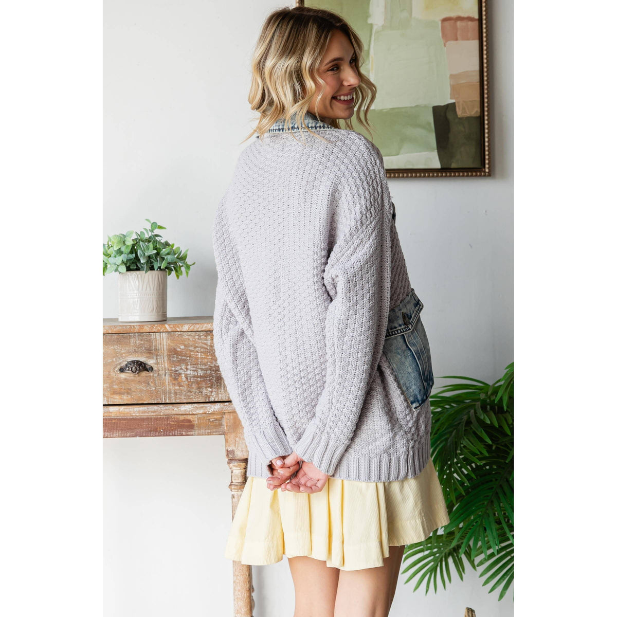 Woman with blonde wavy hair, wearing the Veveret - Knit Body Denim Layerd Jacket: Cream / Denim / L, stands indoors with her back to the camera; a plant, wooden table, and framed art are visible behind her.