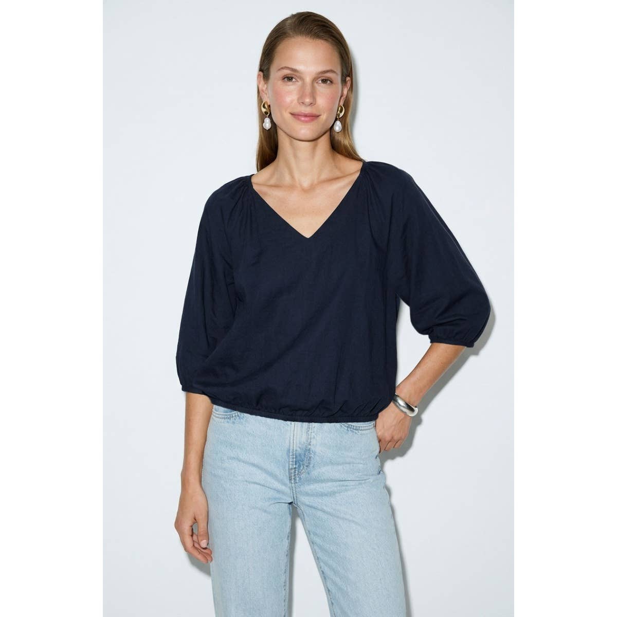 A woman wearing a burgundy Naked Zebra - Elastic Puff Sleeve V-neck Blouse with three-quarter length puff sleeves and light-wash jeans.