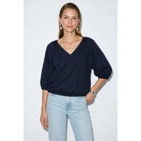 A woman wearing a burgundy Naked Zebra - Elastic Puff Sleeve V-neck Blouse with three-quarter length puff sleeves and light-wash jeans.