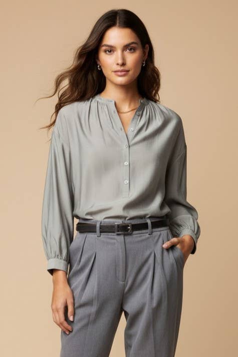 A woman wears an elegant, light gray Naked Zebra - Button-down Blouse With Gathered Neckline tucked into gray pleated trousers with a black belt.