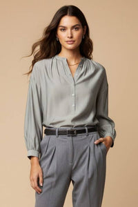 A woman wears an elegant, light gray Naked Zebra - Button-down Blouse With Gathered Neckline tucked into gray pleated trousers with a black belt.