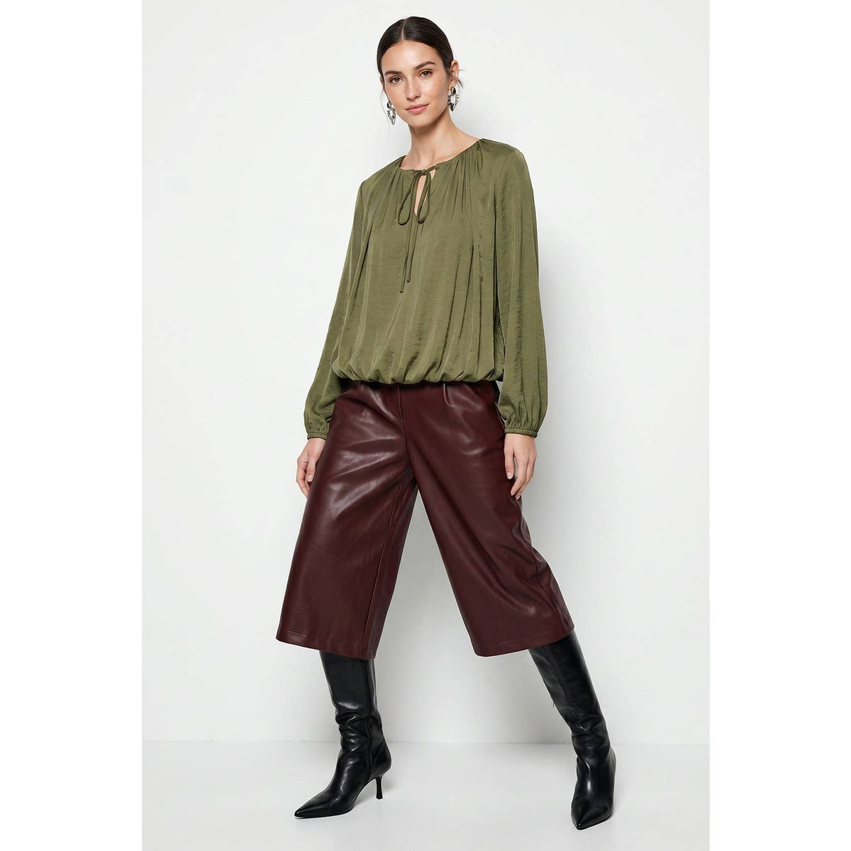 A woman wearing a Naked Zebra - Long Sleeve Draped Bubble Hem Blouse: Wine / L, paired with burgundy cropped pants and black knee-high boots.