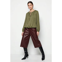 A woman wearing a Naked Zebra - Long Sleeve Draped Bubble Hem Blouse: Wine / L, paired with burgundy cropped pants and black knee-high boots.