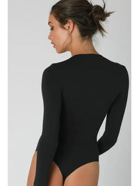 A woman with brown hair styled in a bun wears the High Neck LS Bodysuit—a fitted, long sleeve design perfect for layering. She stands with her back to the camera, turning her head to the side to reveal her profile against a plain background.