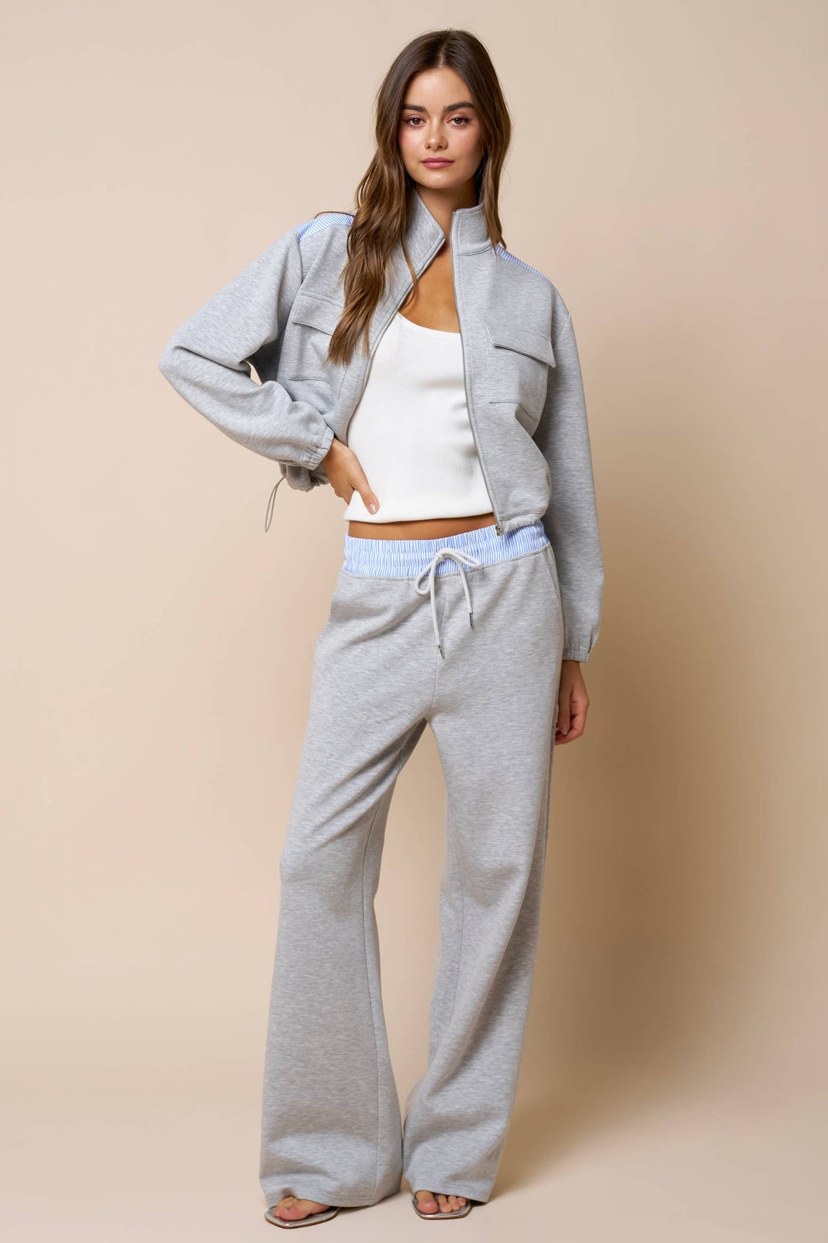 A woman stands against a beige background wearing the Lalavon Color Block Zip Up Mock Neck Sweatshirt in Light Grey and Light Blue (size L) layered over a white tank top, paired with wide-leg pants and her hand in her pocket.