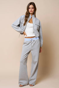 A woman stands against a beige background wearing the Lalavon Color Block Zip Up Mock Neck Sweatshirt in Light Grey and Light Blue (size L) layered over a white tank top, paired with wide-leg pants and her hand in her pocket.