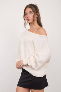 Wearing The Workshop Studio’s Boat Neck Oversized Knit Pullover Sweater in Oatmeal with a black skirt, a woman stands against a plain light background, her hair partially pulled back.