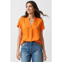 A woman wears a Naked Zebra - Drop Shoulder Regular Blouse in orange, featuring short sleeves, a drop-shoulder design, and a v-neckline, tucked into blue jeans.