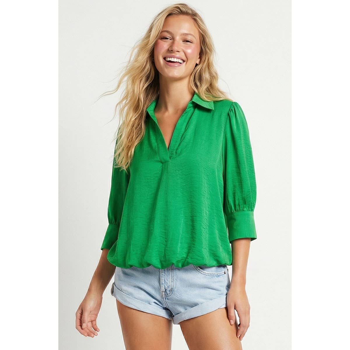 A smiling woman wearing a bright green Naked Zebra - Half Sleeve Hi-lo Bubble Hem Blouse: Wine / L, styled with a collared V-neck and paired with light-wash denim shorts.
