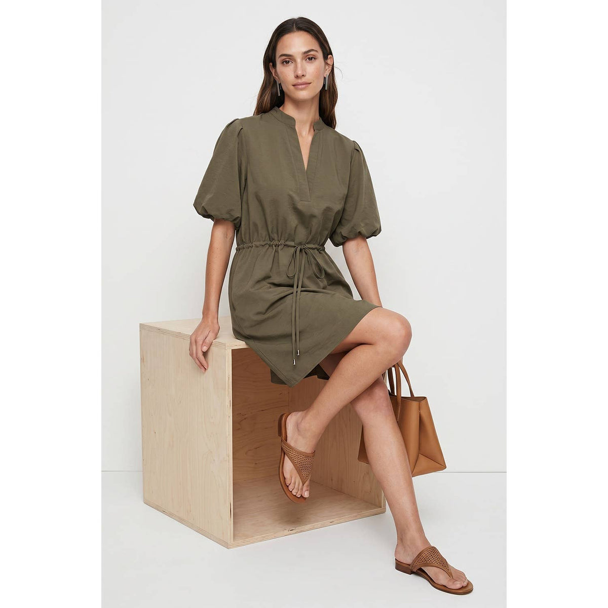 A woman in a navy Naked Zebra - Puff Sleeve Cinched Waist Mini Dress: Navy / L with a cinched waist sits on a wooden cube, wearing brown thong sandals and holding a tan tote bag.